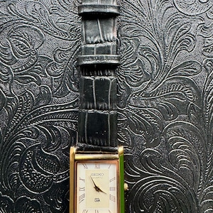 May include: A rectangular gold-toned Seiko Quartz watch with a black leather strap. The watch face is cream-coloured with Roman numerals and the brand name. The strap has a gold-coloured buckle.
