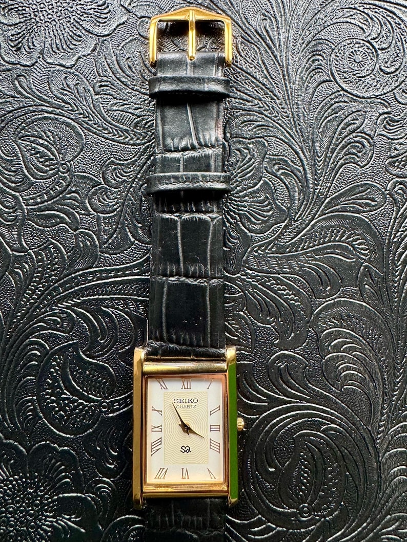 May include: A rectangular gold-toned Seiko Quartz watch with a black leather strap. The watch face is cream-coloured with Roman numerals and the brand name. The strap has a gold-coloured buckle.