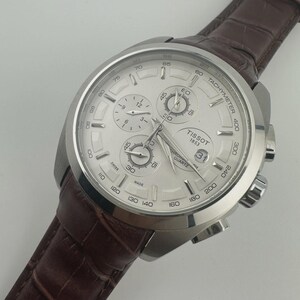 May include: Silver-toned Tissot chronograph watch with a white dial and a brown leather strap. The watch features multiple sub-dials, a date display, and a tachymeter scale. The strap has a textured, crocodile-like pattern.