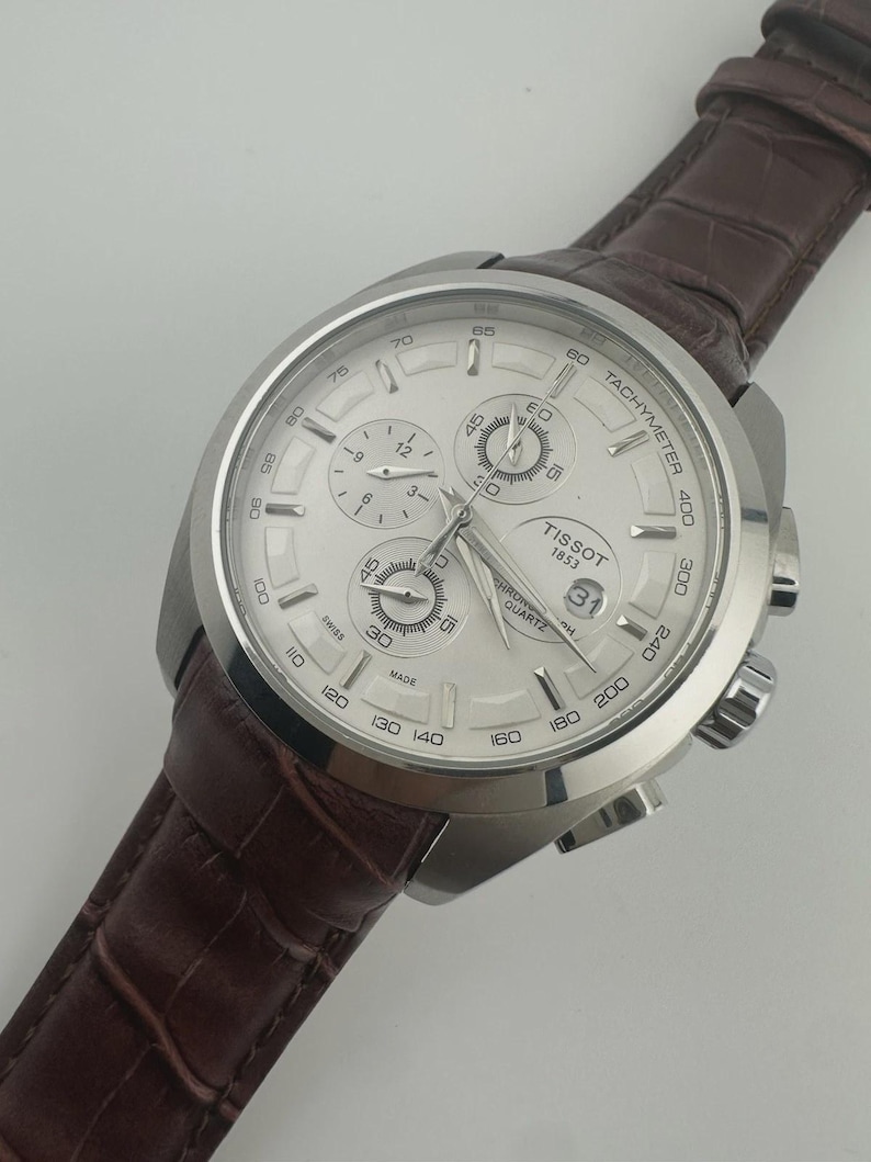 May include: Silver-toned Tissot chronograph watch with a white dial and a brown leather strap. The watch features multiple sub-dials, a date display, and a tachymeter scale. The strap has a textured, crocodile-like pattern.
