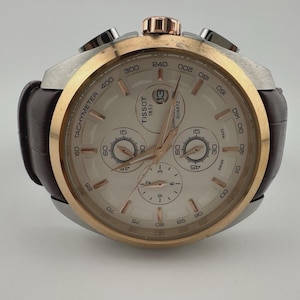 May include: A Tissot quartz chronograph watch with a brown leather strap. The watch features a silver and gold-tone case, a white dial with subdials, and a date display at the 3 o'clock position. The watch has a tachymeter scale and is marked "Tissot 1853".