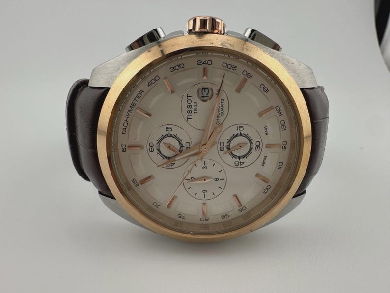 May include: A Tissot quartz chronograph watch with a brown leather strap. The watch features a silver and gold-tone case, a white dial with subdials, and a date display at the 3 o'clock position. The watch has a tachymeter scale and is marked "Tissot 1853".