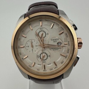 May include: A Tissot chronograph watch with a silver case, gold bezel, and a brown leather strap. The watch face is white with gold accents and three sub-dials. The watch has the text "Tissot 1853 Chronograph Quartz" on the face.