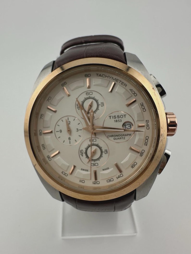 May include: A Tissot chronograph watch with a silver case, gold bezel, and a brown leather strap. The watch face is white with gold accents and three sub-dials. The watch has the text "Tissot 1853 Chronograph Quartz" on the face.