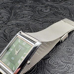 May include: A rectangular Seiko watch with a silver case and a green face featuring Roman numerals. The watch has a mesh silver band. The watch is displayed on a textured black surface.