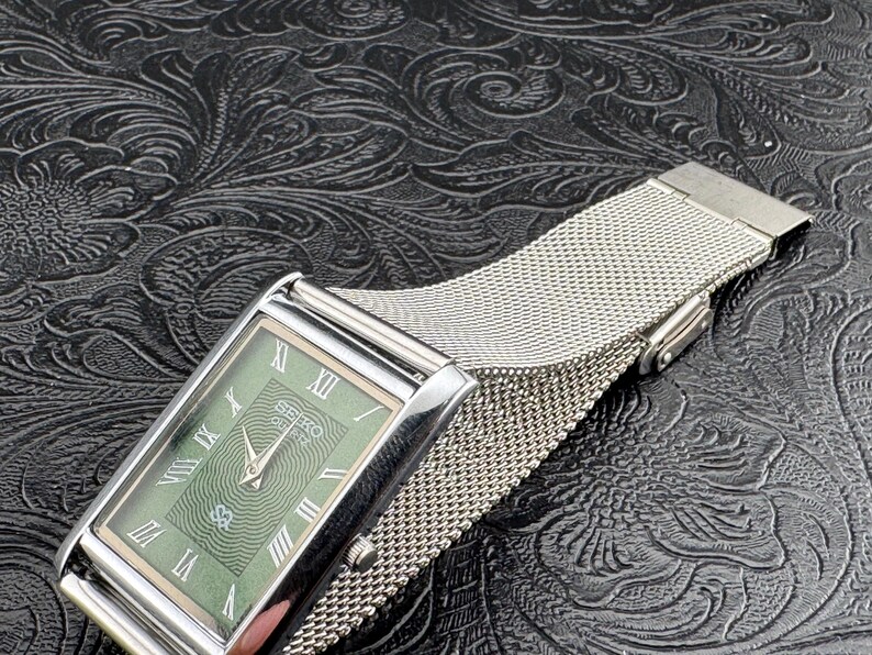 May include: A rectangular Seiko watch with a silver case and a green face featuring Roman numerals. The watch has a mesh silver band. The watch is displayed on a textured black surface.