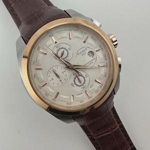 May include: A Tissot chronograph watch with a silver face, gold-tone bezel, and brown leather strap. The watch features multiple sub-dials, a date window, and a tachymeter scale. The watch is a stylish accessory.