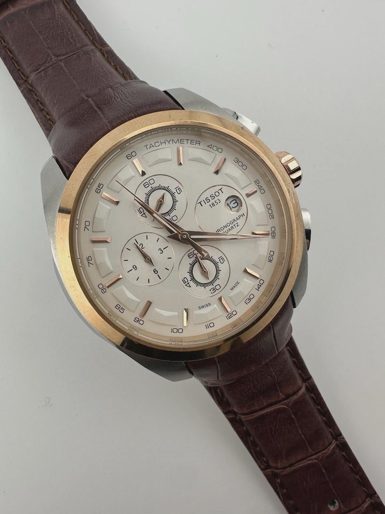 May include: A Tissot chronograph watch with a silver face, gold-tone bezel, and brown leather strap. The watch features multiple sub-dials, a date window, and a tachymeter scale. The watch is a stylish accessory.