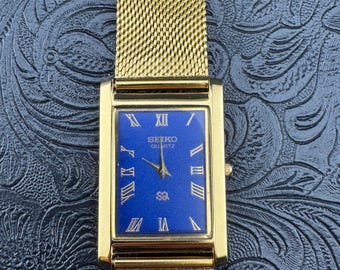 May include: A gold-toned Seiko Quartz watch with a rectangular face and a mesh band. The watch face is a vibrant blue with gold Roman numerals and hands. The watch is displayed on a textured, dark blue surface.
