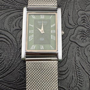 May include: A rectangular Seiko Quartz watch with a silver mesh band. The watch face is a muted green with Roman numerals and a textured center. The watch hands point to 10:09. The watch has a silver bezel and a small crown.
