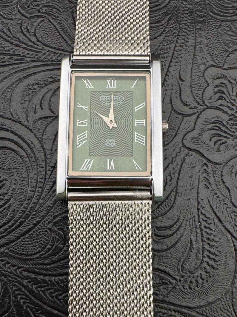 May include: A rectangular Seiko Quartz watch with a silver mesh band. The watch face is a muted green with Roman numerals and a textured center. The watch hands point to 10:09. The watch has a silver bezel and a small crown.