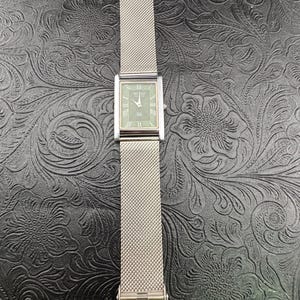 May include: A rectangular Seiko watch with a silver mesh band. The watch face is a muted green with silver hands and Roman numeral hour markers. The band is secured with a clasp that reads "SEIKO".