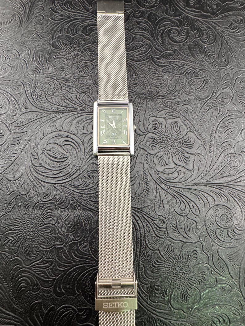 May include: A rectangular Seiko watch with a silver mesh band. The watch face is a muted green with silver hands and Roman numeral hour markers. The band is secured with a clasp that reads "SEIKO".