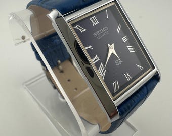 May include: A rectangular Seiko Quartz watch with a dark blue face and silver-tone case. The watch features Roman numeral hour markers and a blue leather band. The watch is displayed on a clear acrylic stand.