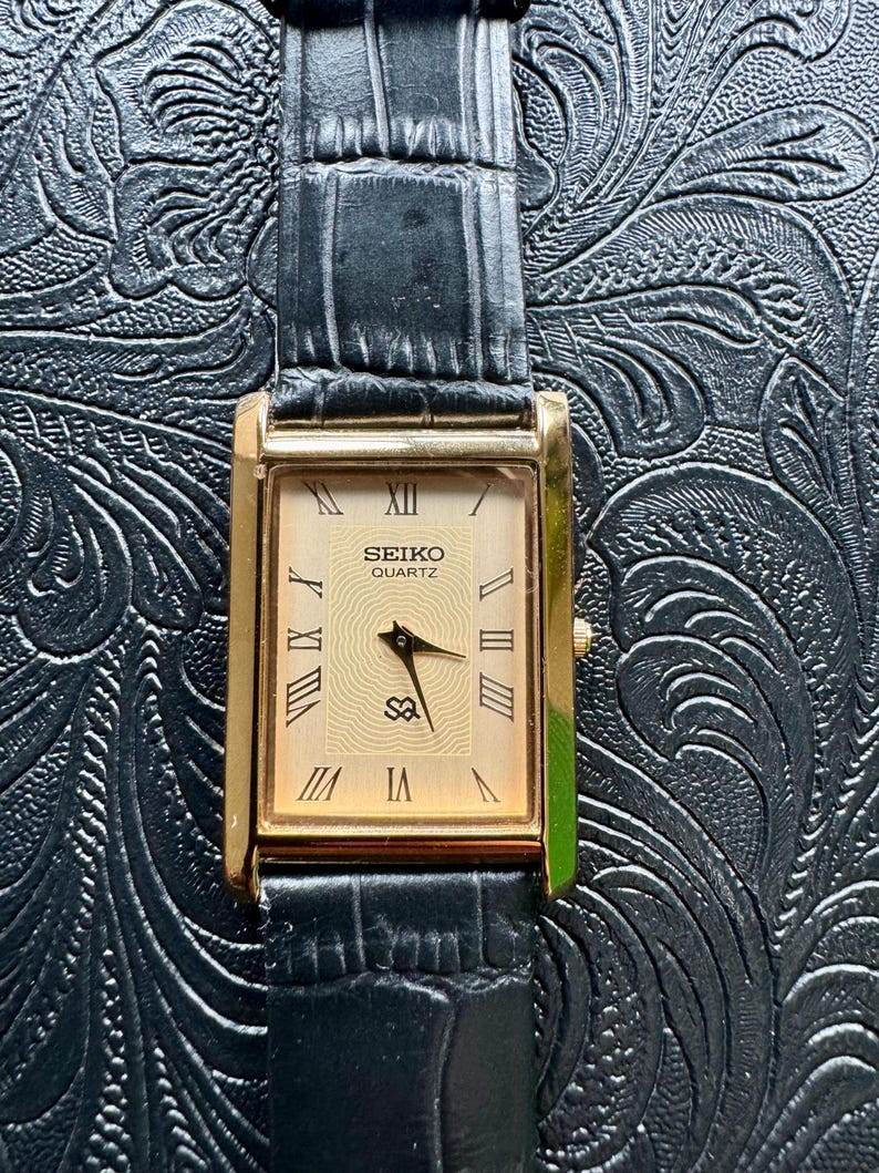 May include: A rectangular Seiko Quartz watch with a gold-tone case and a cream-coloured face. The face features Roman numeral hour markers and black hands. The watch has a black leather strap with a crocodile pattern.