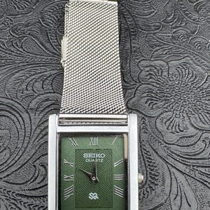 May include: A rectangular Seiko Quartz watch with a silver-tone case and a mesh band. The watch face is green with Roman numerals and the Seiko logo. The watch has a silver crown.