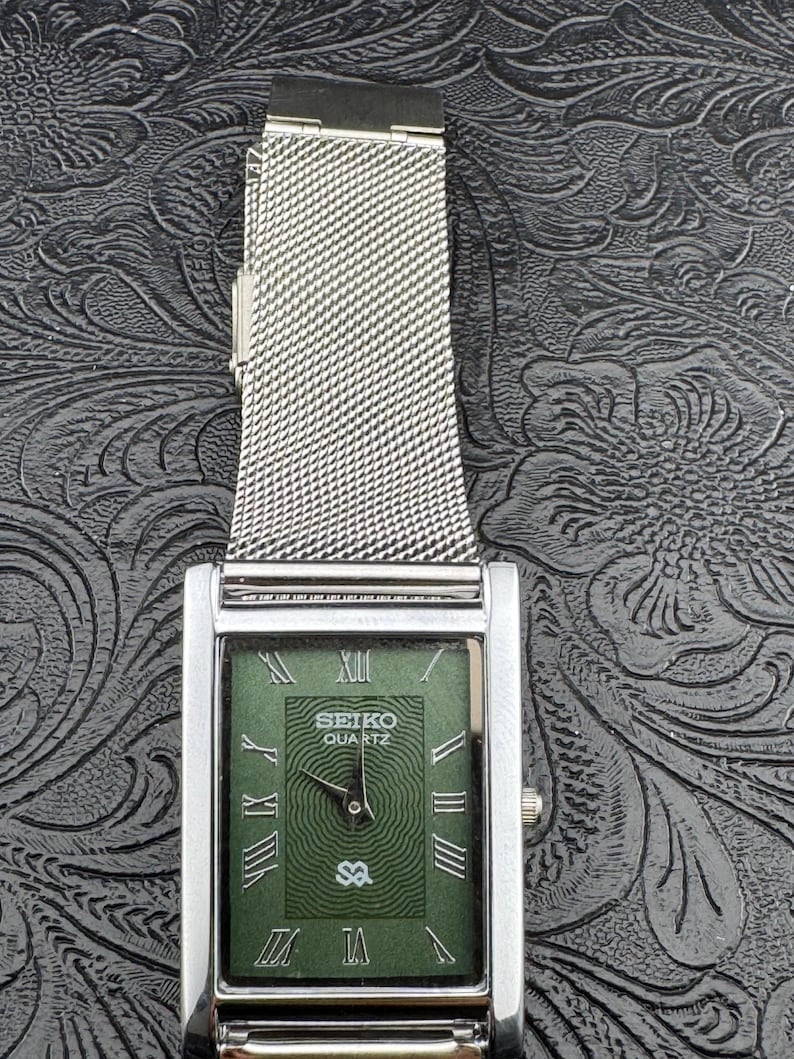 May include: A rectangular Seiko Quartz watch with a silver-tone case and a mesh band. The watch face is green with Roman numerals and the Seiko logo. The watch has a silver crown.