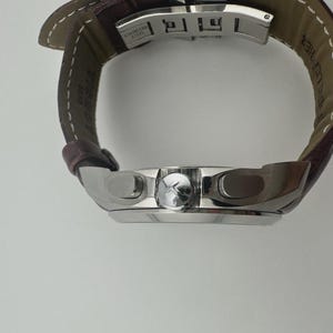 May include: A silver wristwatch with a brown leather band. The watch face has a silver bezel and a dark dial. The band is stitched with white thread. The watch is a luxury item.