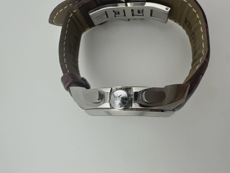 May include: A silver wristwatch with a brown leather band. The watch face has a silver bezel and a dark dial. The band is stitched with white thread. The watch is a luxury item.