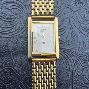 May include: A gold-toned Seiko Quartz watch with a rectangular face and a mesh-style band. The watch face is white with Roman numerals and the Seiko logo. The watch is displayed on a dark, textured surface.