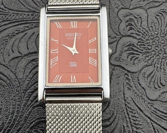 May include: A rectangular Seiko Quartz watch with a silver mesh band. The watch face is a salmon color with Roman numerals and the Seiko logo. The watch has a silver case and a mesh band.