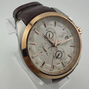 May include: A silver and gold-tone Tissot chronograph watch with a brown leather strap. The watch face is light-colored with multiple sub-dials and rose gold accents. The watch is displayed on a clear stand.