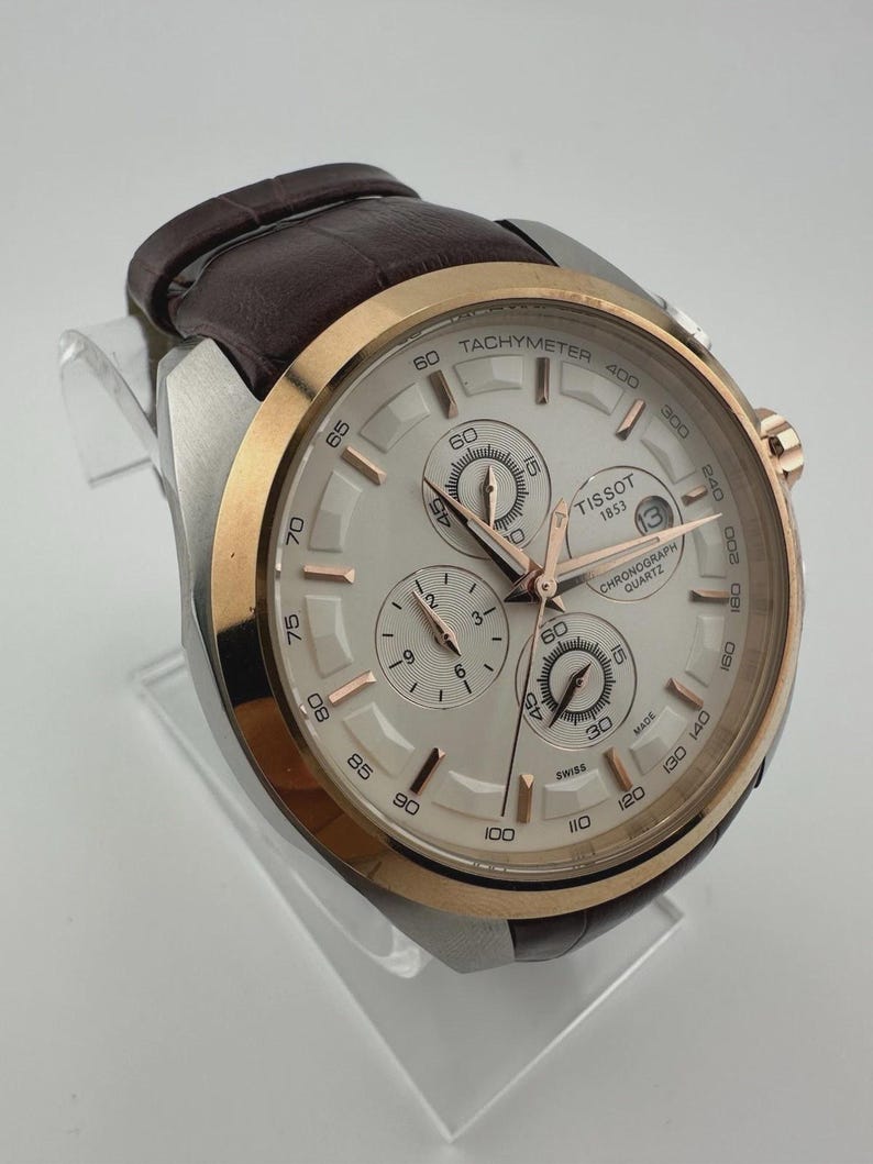May include: A silver and gold-tone Tissot chronograph watch with a brown leather strap. The watch face is light-colored with multiple sub-dials and rose gold accents. The watch is displayed on a clear stand.