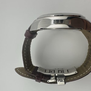 May include: A silver-toned wristwatch with a brown leather band. The watch face is not visible, but the stainless steel case and clasp are prominent. The band is stitched with white thread and marked "GENUINE LEATHER".