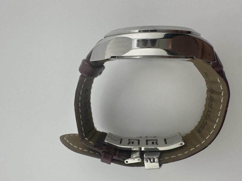 May include: A silver-toned wristwatch with a brown leather band. The watch face is not visible, but the stainless steel case and clasp are prominent. The band is stitched with white thread and marked "GENUINE LEATHER".