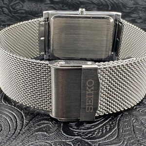 May include: A silver-toned Seiko watch with a rectangular face and a mesh band. The clasp is engraved with the Seiko brand name. The watch is displayed on a textured black surface.