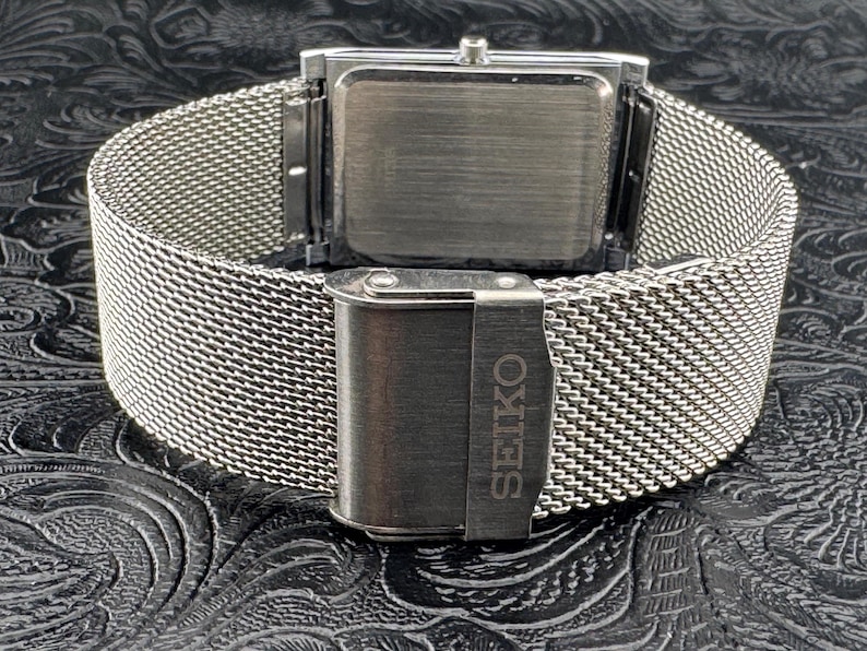 May include: A silver-toned Seiko watch with a rectangular face and a mesh band. The clasp is engraved with the Seiko brand name. The watch is displayed on a textured black surface.