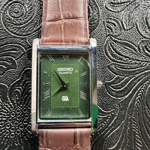 May include: A rectangular Seiko Quartz watch with a green face and Roman numerals. The watch has a brown leather band with a crocodile pattern. The watch face has the Seiko Quartz logo and a unique design.
