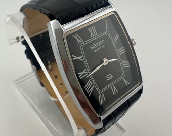 Peut inclure: Une montre Seiko Quartz rectangulaire avec un cadran noir et un boîtier argenté. La montre présente des chiffres romains pour les heures et un bracelet en cuir noir. La montre est présentée sur un présentoir en acrylique transparent.