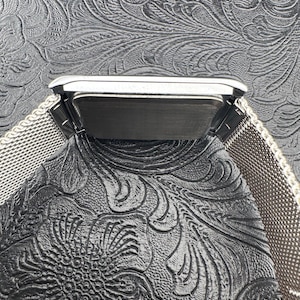 May include: A silver-toned rectangular watch with a mesh band. The watch face is dark, and the band is made of a fine, woven metal. The watch is set against a textured, dark background.