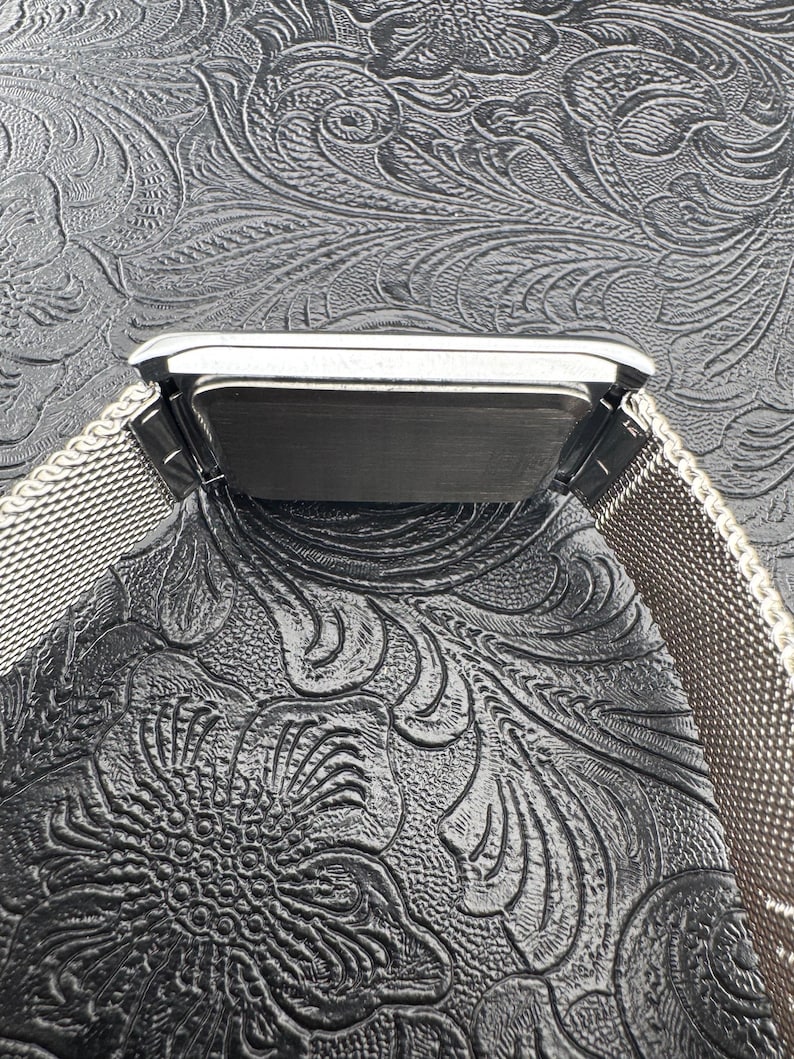 May include: A silver-toned rectangular watch with a mesh band. The watch face is dark, and the band is made of a fine, woven metal. The watch is set against a textured, dark background.