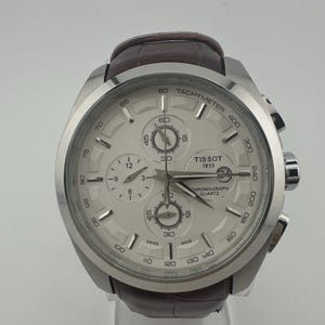 May include: A silver Tissot chronograph quartz watch with a white face and brown leather strap. The watch features multiple subdials, a tachymeter scale, and the Tissot 1853 logo. The watch is displayed on a clear stand.