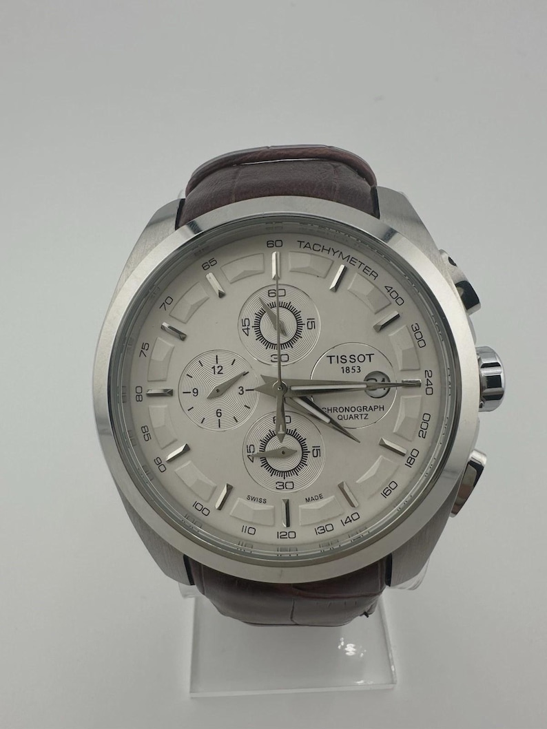 May include: A silver Tissot chronograph quartz watch with a white face and brown leather strap. The watch features multiple subdials, a tachymeter scale, and the Tissot 1853 logo. The watch is displayed on a clear stand.