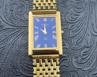 May include: Gold-toned Seiko Quartz watch with a rectangular face and a blue dial. The watch features Roman numeral hour markers and a gold-toned mesh band. The watch is a classic design.