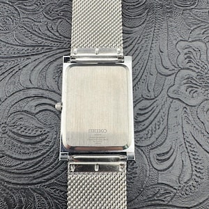 May include: A silver-toned Seiko quartz watch with a rectangular face and a mesh band. The watch face is silver, and the band is a fine, woven mesh. The back of the watch is visible, with the Seiko logo and other text.