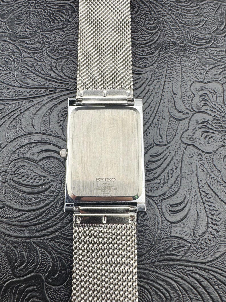 May include: A silver-toned Seiko quartz watch with a rectangular face and a mesh band. The watch face is silver, and the band is a fine, woven mesh. The back of the watch is visible, with the Seiko logo and other text.