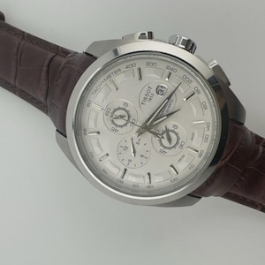 May include: A silver Tissot chronograph watch with a white face and a brown leather band. The watch features multiple sub-dials and a tachymeter scale. The band is a dark brown color with a textured, crocodile-like pattern.
