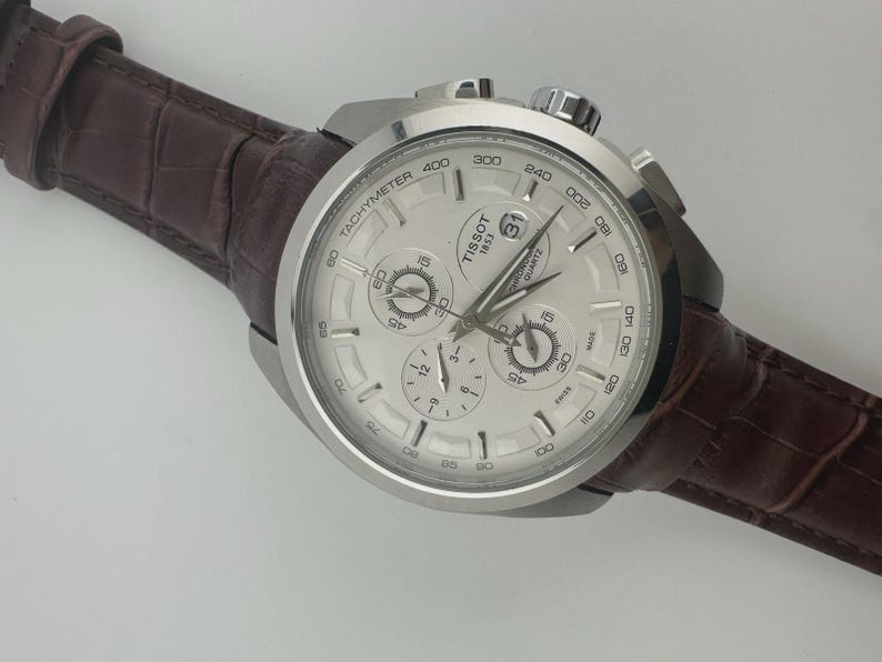 May include: A silver Tissot chronograph watch with a white face and a brown leather band. The watch features multiple sub-dials and a tachymeter scale. The band is a dark brown color with a textured, crocodile-like pattern.