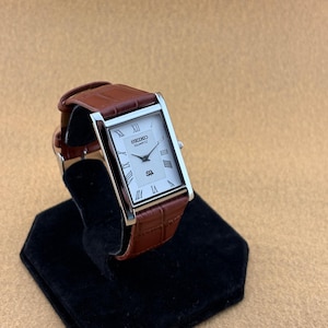 May include: A rectangular Seiko Quartz wristwatch featuring a white dial with black Roman numeral hour markers, a silver-tone metal case, and a brown leather strap with a textured crocodile-embossed pattern and a silver-tone buckle closure.