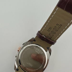 May include: A Tissot watch with a brown leather strap. The watch face is stainless steel with rose gold accents. The back of the watch is visible, displaying the brand name, "Tissot 1853", and "Sapphire Crystal". The strap size is 22.