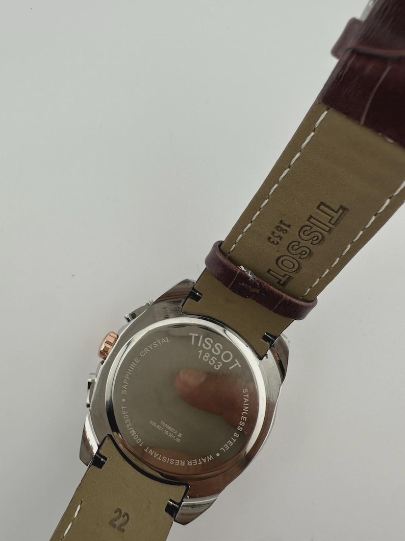 May include: A Tissot watch with a brown leather strap. The watch face is stainless steel with rose gold accents. The back of the watch is visible, displaying the brand name, "Tissot 1853", and "Sapphire Crystal". The strap size is 22.