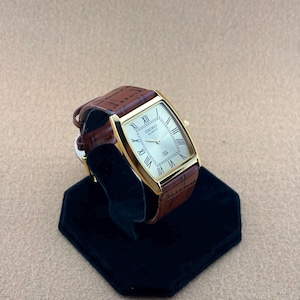 May include: A gold-tone rectangular wristwatch with a champagne-colored dial featuring black Roman numeral hour markers. The watch face displays the text SEIKO QUARTZ and a small SQ logo. It is fitted with a brown leather strap with a textured finish.