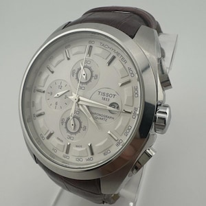 May include: A silver Tissot chronograph watch with a white dial and brown leather strap. The watch features multiple sub-dials, a tachymeter scale, and the text "TISSOT 1853 CHRONOGRAPH QUARTZ". The watch is displayed on a clear stand.