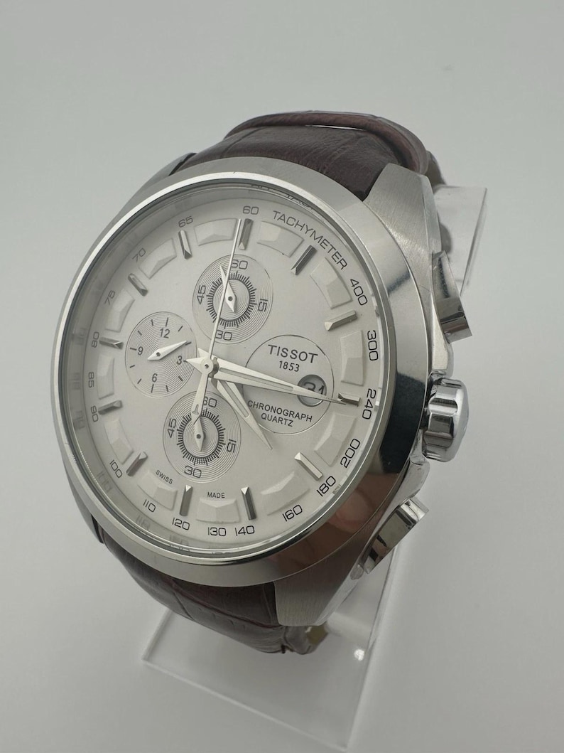 May include: A silver Tissot chronograph watch with a white dial and brown leather strap. The watch features multiple sub-dials, a tachymeter scale, and the text "TISSOT 1853 CHRONOGRAPH QUARTZ". The watch is displayed on a clear stand.