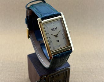 May include: A rectangular Seiko Quartz watch with a gold-tone case and a champagne-colored dial featuring Roman numeral hour markers. The watch has a dark green leather strap with a textured finish and visible green stitching along the edges.