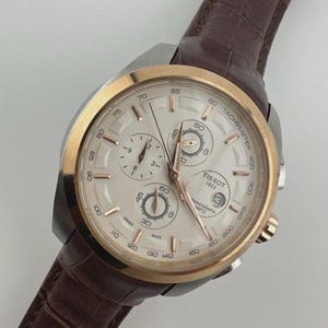 May include: A Tissot chronograph quartz watch with a silver face, gold bezel, and brown leather strap. The watch face displays multiple sub-dials and the date. The watch is angled, showcasing the details of the dial and strap.